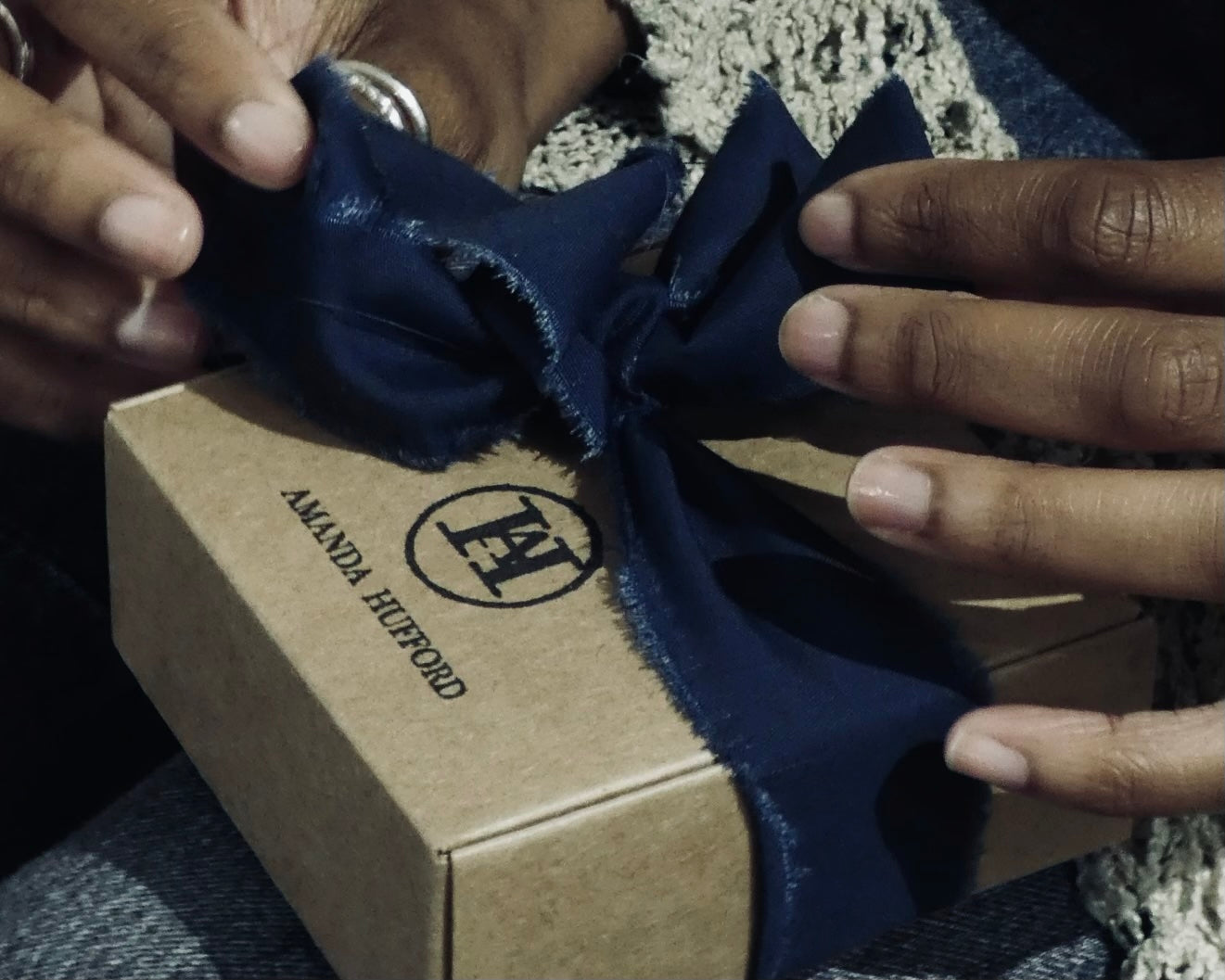 Close-up of hands tying the bow on Amanda Hufford Studio's signature branded packaging — a recycled Kraft box with a stamped logo and recycled navy indigo blue fabric ribbon.
