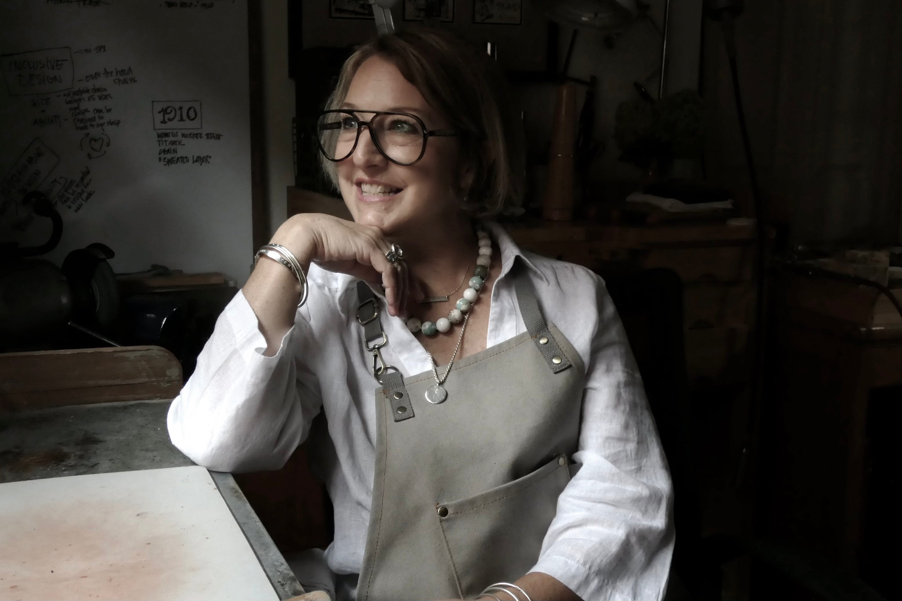 Amanda Hufford sitting at her jewelry studio bench, wearing a workshop apron and handmade Amanda Hufford Studio jewelry designs